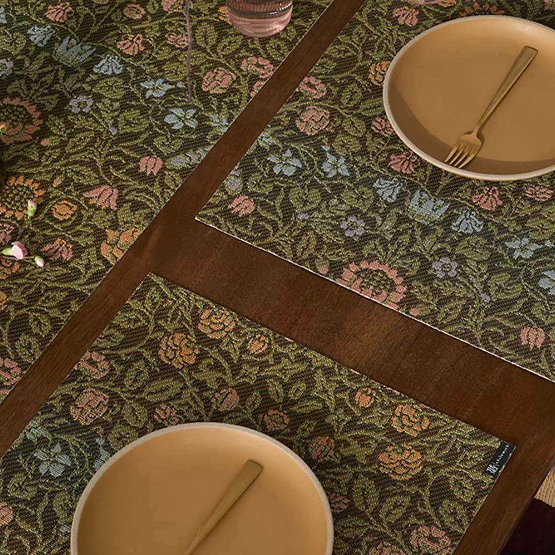 A woven placemat with a floral design in brown, green, and pink colors, featuring a small label in the bottom right corner.