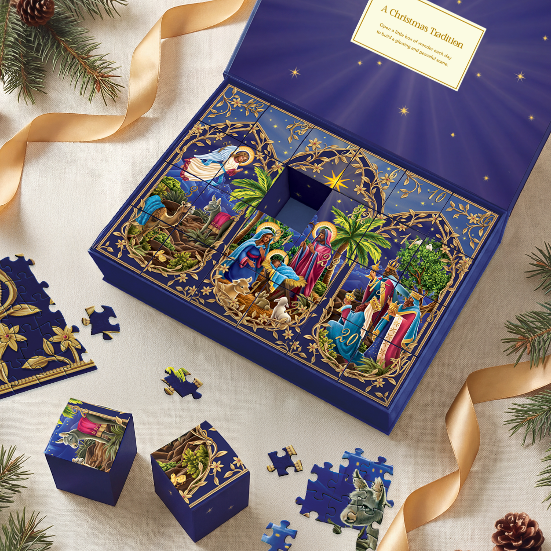 A Christmas-themed advent-style gift box set displayed on a light fabric surface with pine branches, gold ribbon, and puzzle pieces around it. The main open box is deep blue with gold star details and illustrated nativity scenes on the front panels.