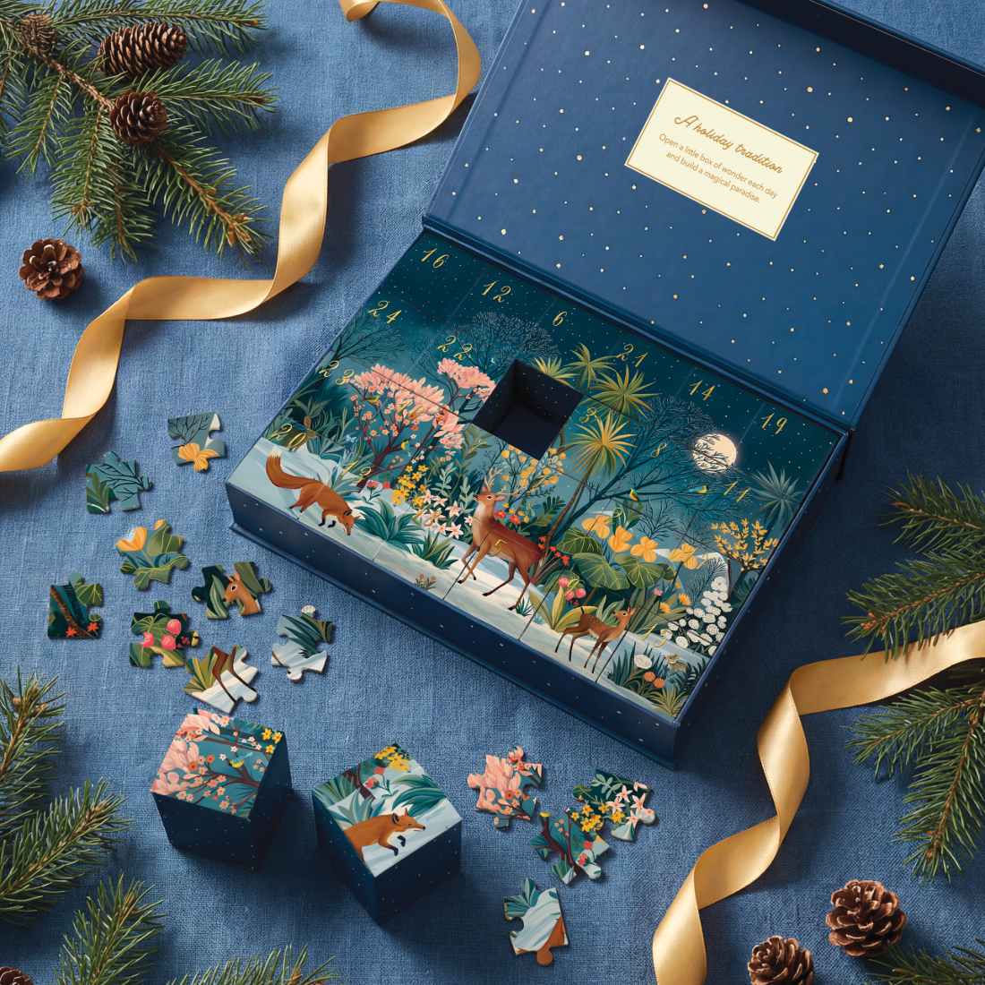 A decorative illustrated advent box set displayed on a blue fabric surface with pine branches, gold ribbon, and pinecones. The open box shows a whimsical forest scene with animals and numbered compartments, and several matching puzzle pieces and small boxes are arranged around it.