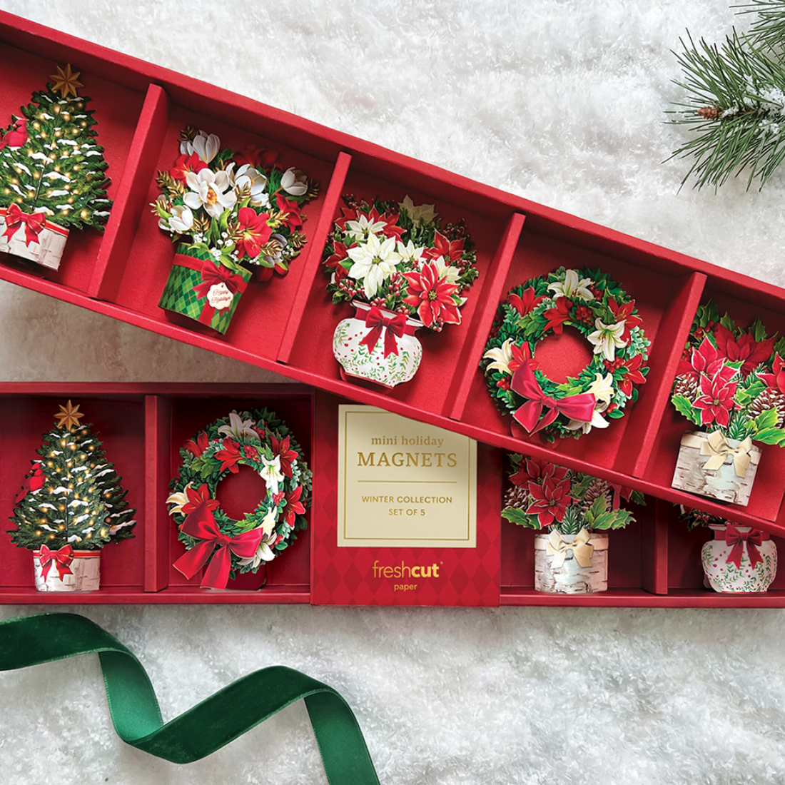 A red gift box filled with small Christmas-themed magnets arranged in compartments, including a tree, wreaths, flowers, and holiday motifs. A gold label reads 'mini holiday MAGNETS winter collection set of 5' and the brand 'freshcut' is visible.