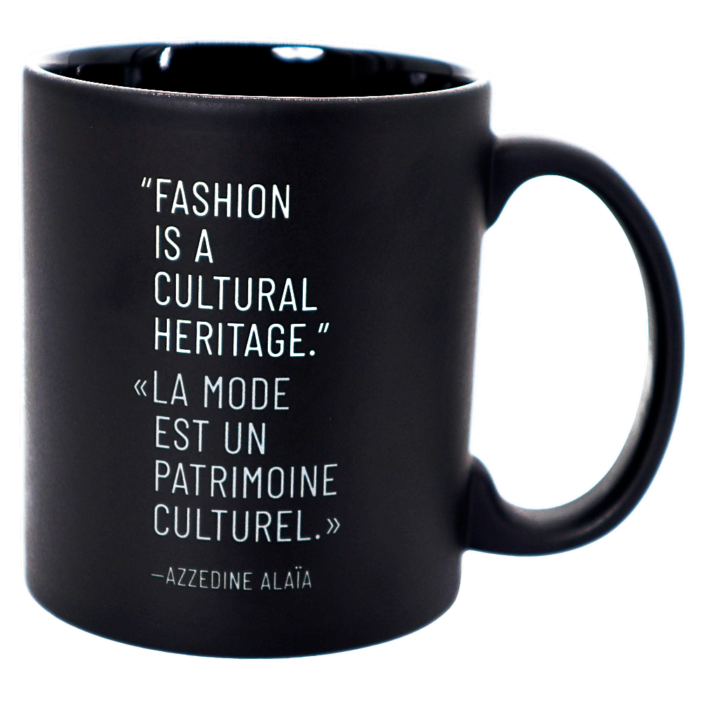 A matte black ceramic mug with white printed text featuring a quote by Azzedine Alaia, shown on a plain white background.