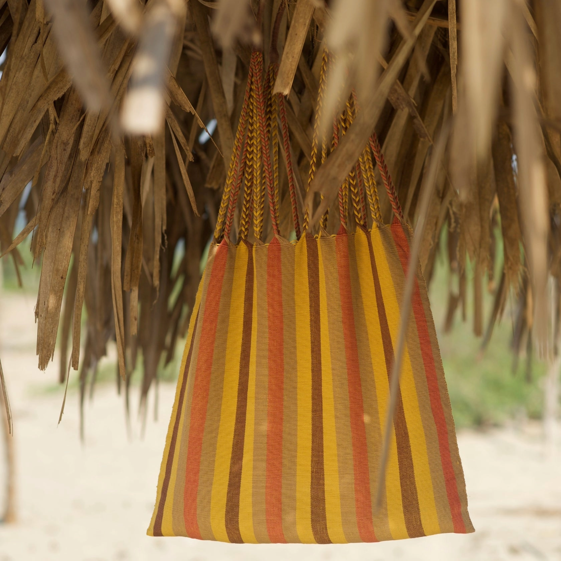 A woven hanging bag with vertical stripes in yellow, orange, and brown, suspended from a thatched roof outdoors.