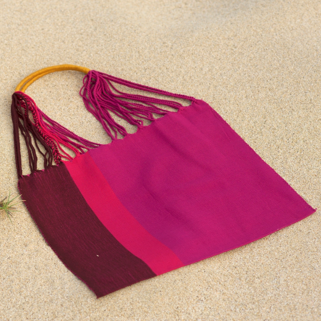A woven shoulder bag made from magenta and burgundy wool with a mustard yellow handle, laid flat on a sandy beige background.