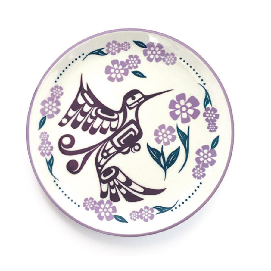 A round porcelain plate with a contemporary Indigenous design featuring a hummingbird and floral patterns in purple and green hues on a white background.