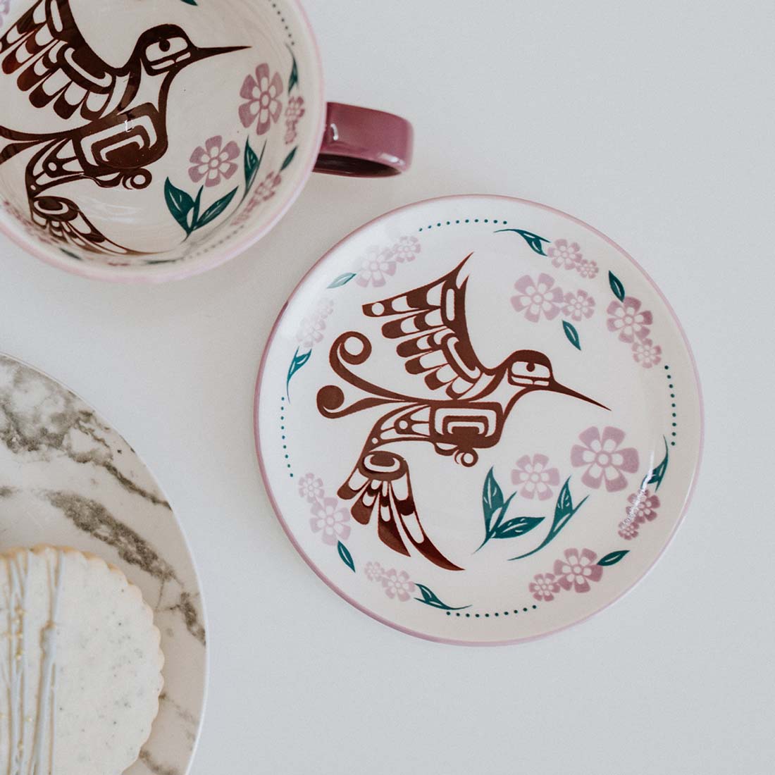 A round porcelain plate with a contemporary Indigenous design featuring a hummingbird and floral patterns in purple and green hues on a white background.