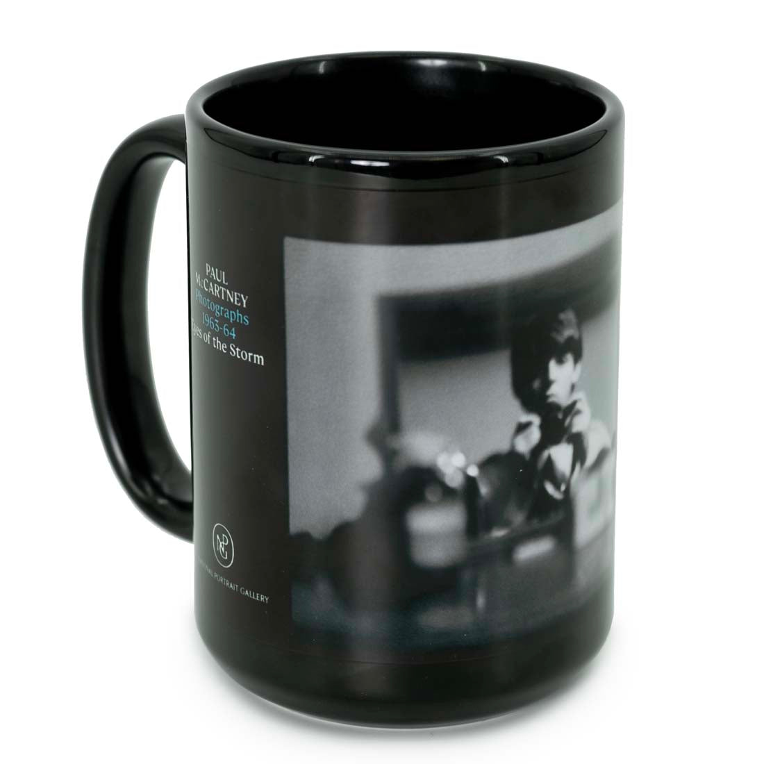 A black ceramic mug with a monochrome photograph of Paul McCartney and text related to the de Young museum exhibition.