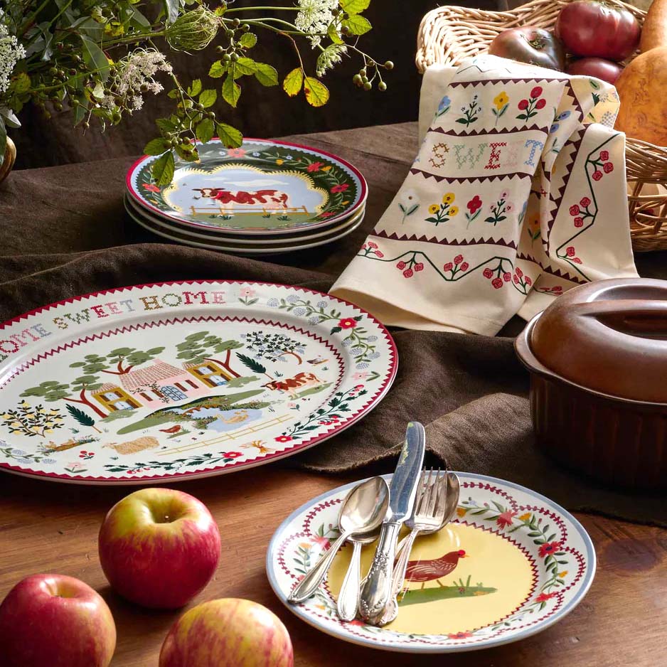 Decorative plates with rural scenes, a towel, apples, and a pot on a wooden table.