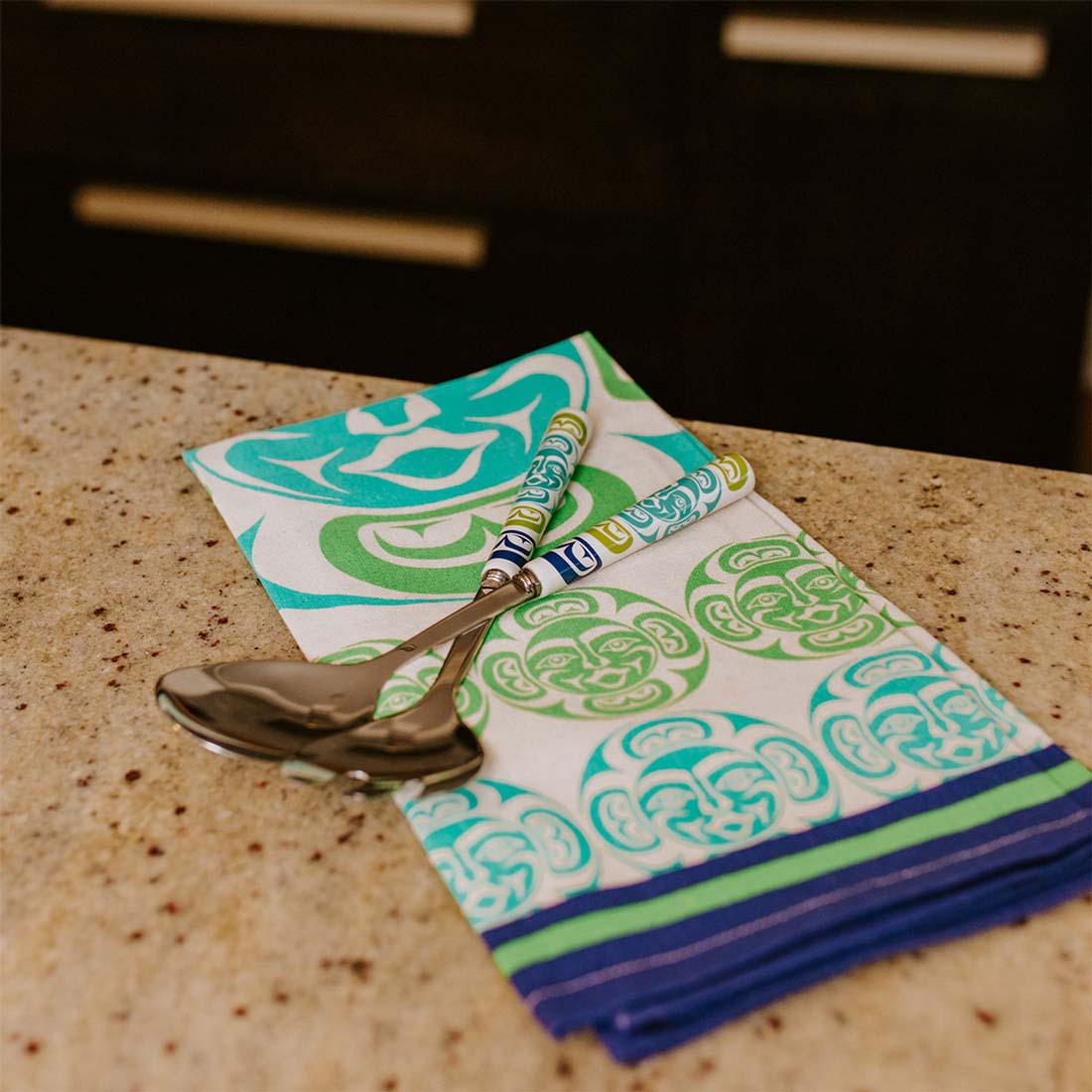 A multicolored cotton tea towel featuring a geometric pattern with face motifs in blue and green on a natural beige cotton, with a blue border.